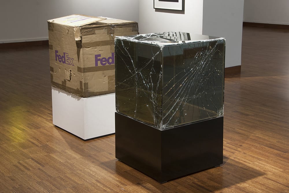 two boxes on plinths in a museum. one box is a large fedex cardboard box, and the other is a box almost the same size made of glass, with many large cracks across the surface.