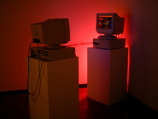 two crt monitors on plinths. one has red text on the monitor and the two computers are connected by a cable. they are lit with red lights.